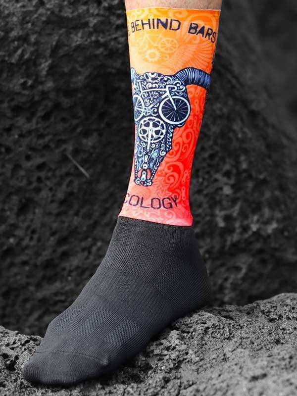 Cycling sock with bull skull and bike design, vibrant orange and red, 'Behind Bars' text, rugged terrain background