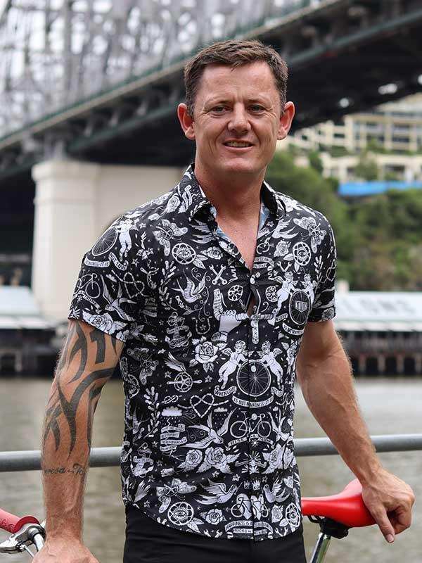 Man wearing black and white cycling-themed shirt with bike graphics, standing by river with bridge background