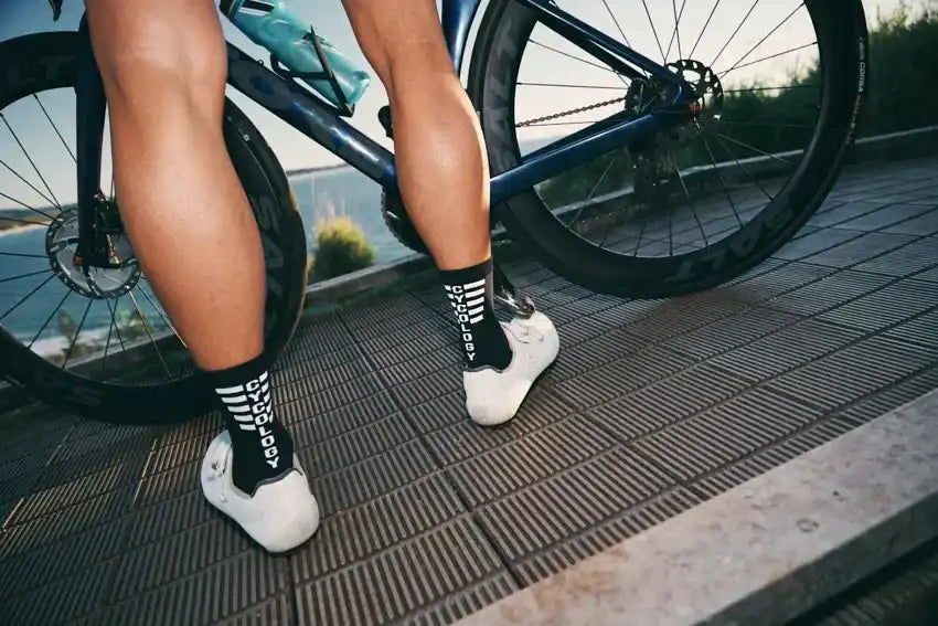 Cyclist wearing black socks with white stripes and white cycling shoes beside a blue bike on a paved path.