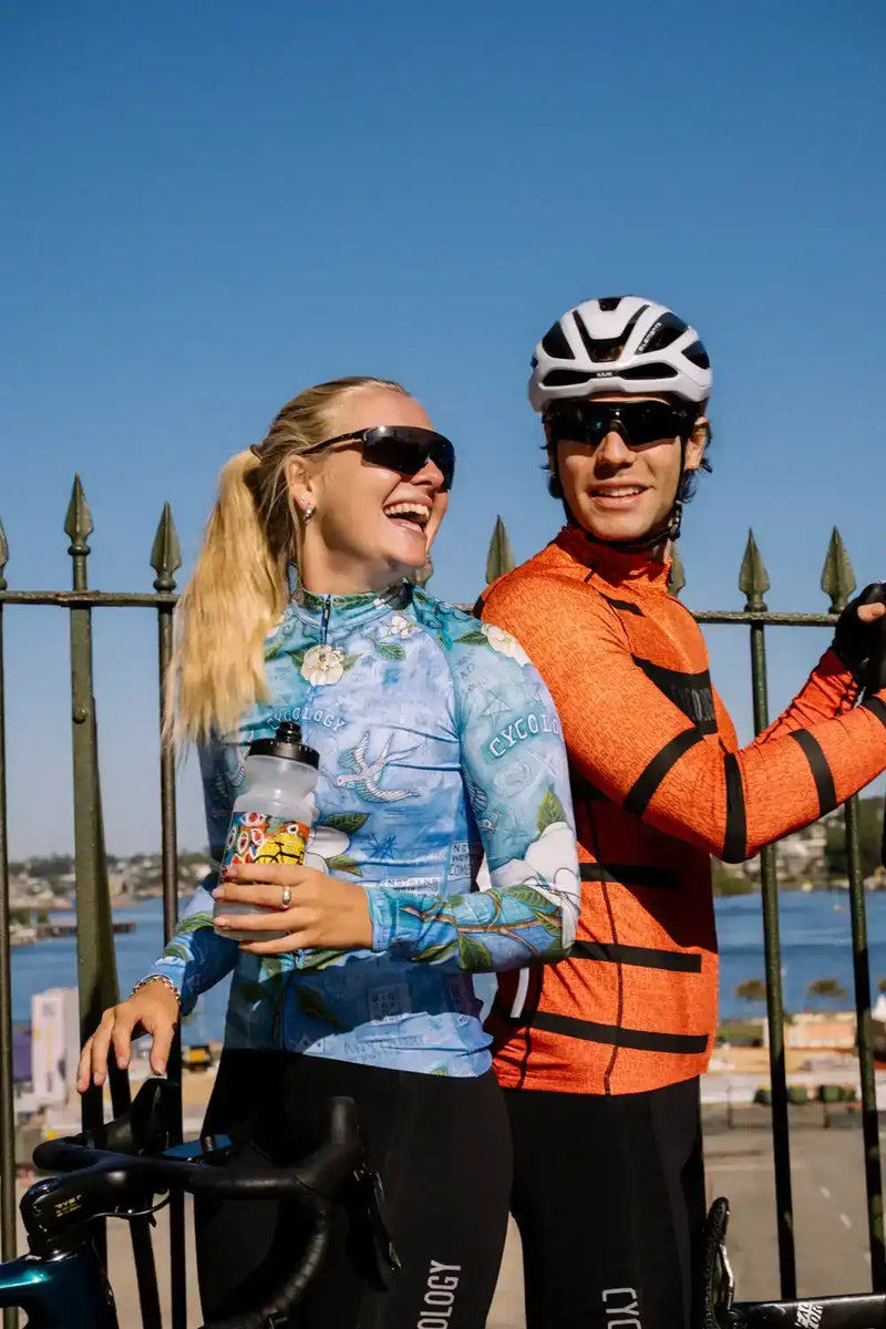 Man and woman in colorful cycling jerseys smiling by waterfront, wearing helmets and sunglasses