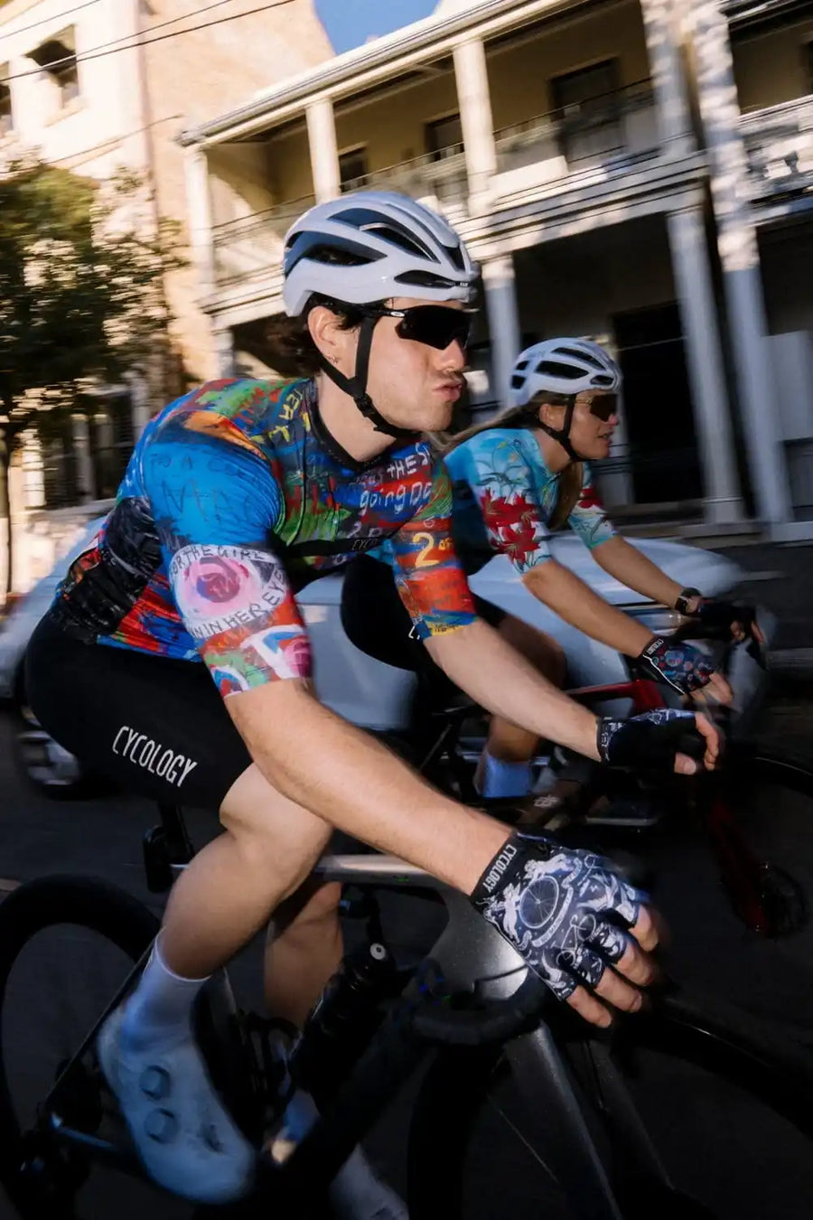 Man and woman cycling in colorful Cycology jerseys on city streets, wearing helmets and gloves