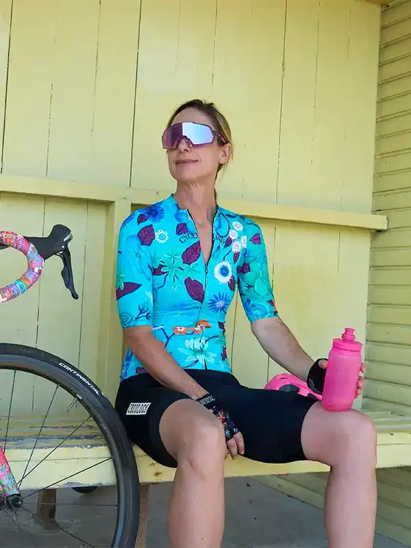 Woman in floral cycling jersey and sunglasses holding pink water bottle, seated beside bike