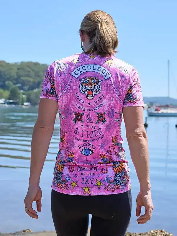Woman wearing vibrant pink cycling jersey with tiger graphic and 'Cycology' back print, standing by water
