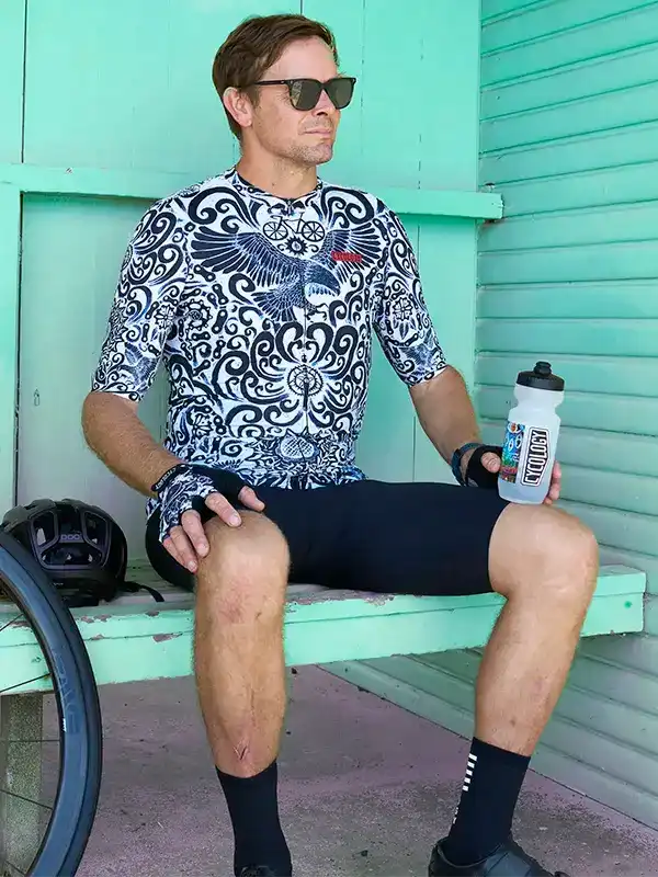 Man in patterned cycling jersey and shorts holding water bottle, seated beside bike near mint wall