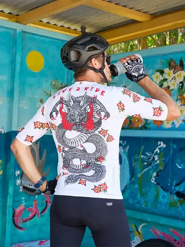 Man wearing dragon-print cycling jersey and helmet, drinking water at colorful mural backdrop