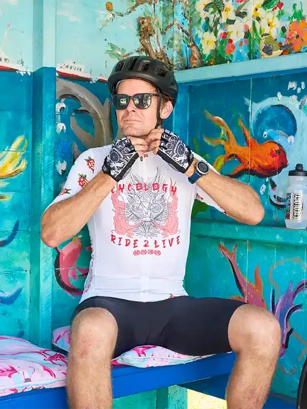 Man in cycling jersey and helmet adjusting gloves, seated against colorful mural backdrop
