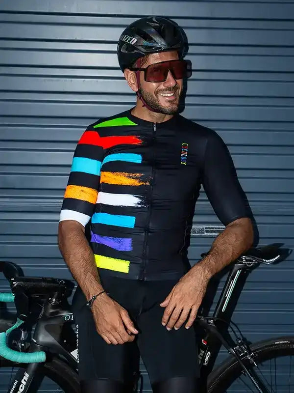 Man in colorful striped cycling jersey and helmet, standing with road bike against gray shutter