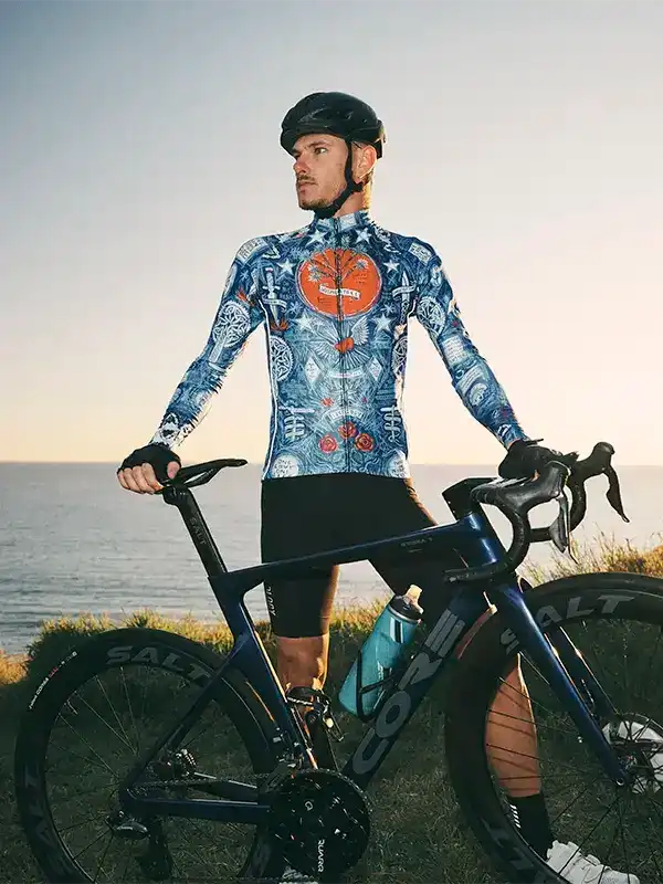 Man in blue patterned cycling jersey and black shorts standing with road bike by ocean at sunset
