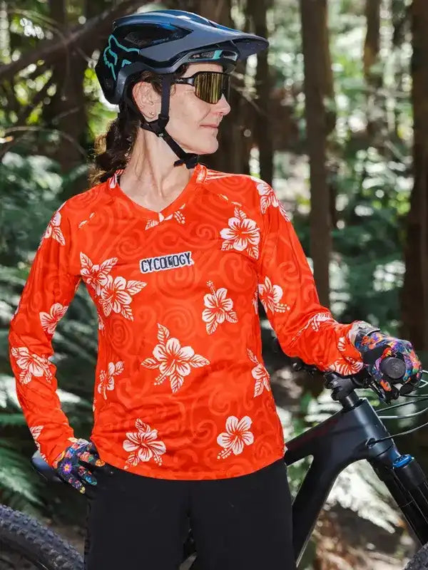 Woman in orange floral cycling jersey with 'Cycology' logo, helmet, and gloves, standing with mountain bike in forest