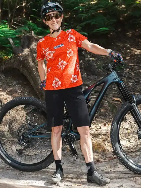 Woman in vibrant red floral cycling jersey and black shorts, standing with mountain bike on forest trail