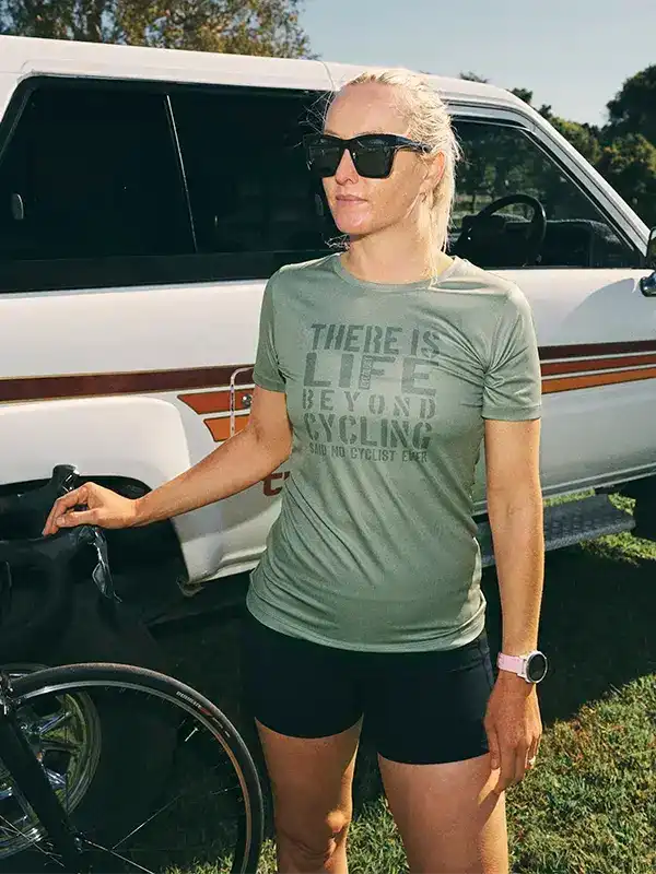 Woman in olive cycling t-shirt with 'There Is Life Beyond Cycling' slogan, standing by bike and van outdoors