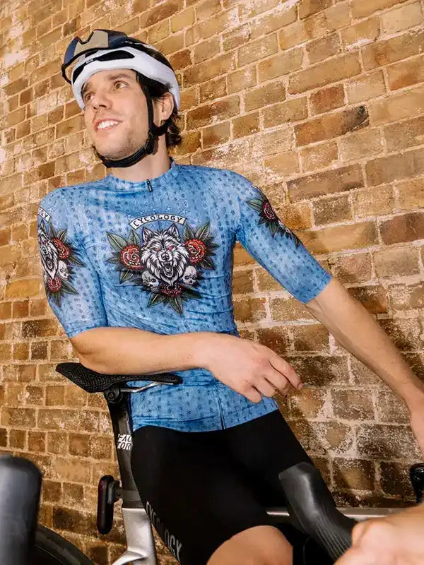 Man wearing blue cycling jersey with wolf and rose graphic, black shorts, helmet, on stationary bike against brick wall