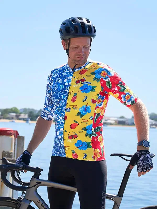 Man in colorful tropical cycling jersey and black bib shorts, standing by bike near water, wearing helmet and gloves