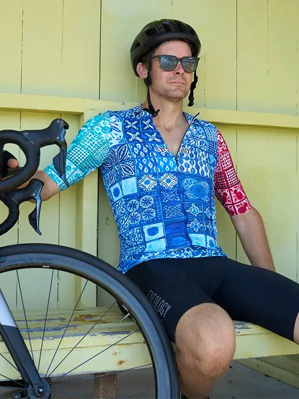 Man in blue patterned cycling jersey and black shorts, leaning on bike near yellow wall