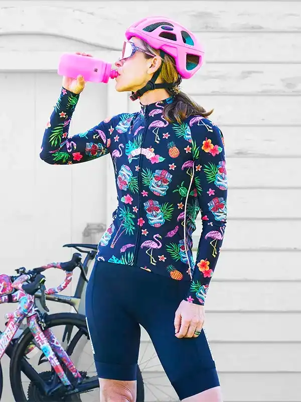 Woman in tropical print cycling jersey and pink helmet drinking from water bottle, bike nearby