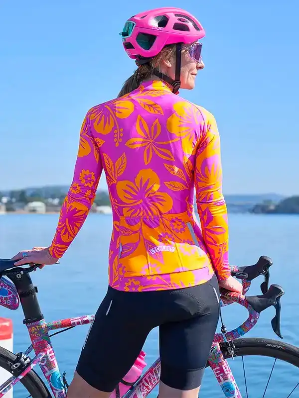 Woman in vibrant floral cycling jersey and pink helmet by water, holding bike