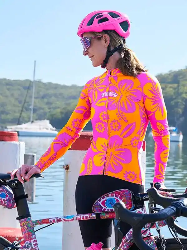 Woman in vibrant pink and orange floral cycling jersey with helmet, standing by bike near water