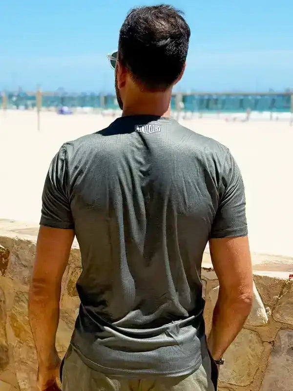 Man wearing gray cycling jersey with 'CYCOLOGY' logo, standing on beach overlooking ocean