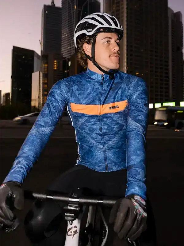 Man in blue patterned cycling jersey with orange stripe, wearing helmet, on bike in city at dusk