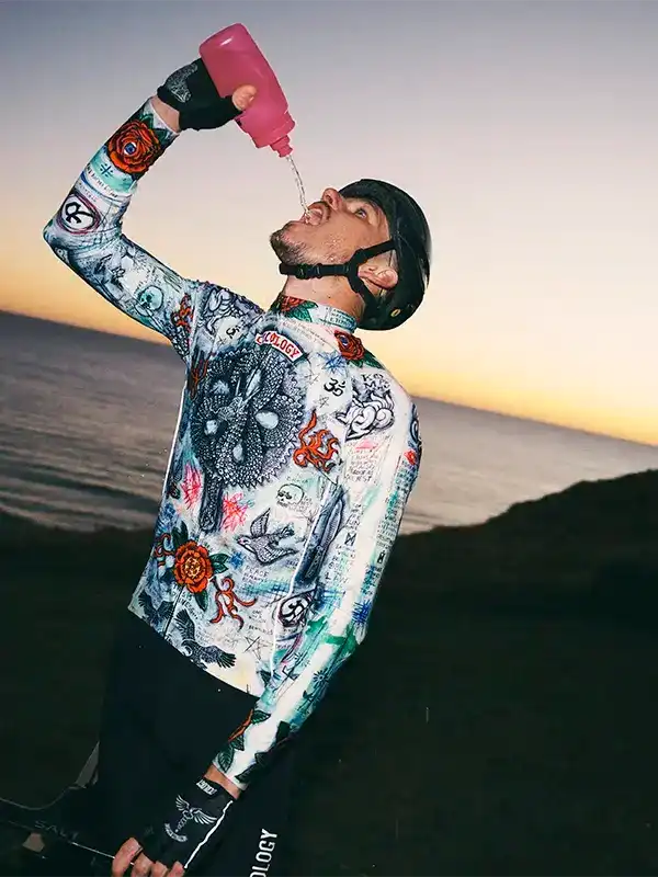 Cyclist drinking from pink bottle, wearing tattoo-print cycling jersey at sunset
