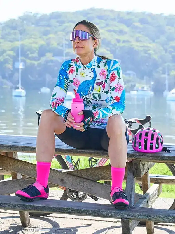 Woman in floral cycling jersey and pink socks holding water bottle, helmet beside her, by waterfront