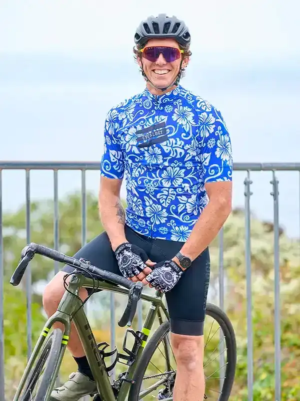 Man in blue floral cycling jersey and black shorts, smiling with road bike outdoors