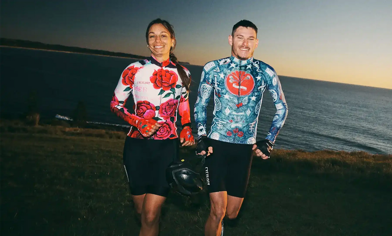Man and woman in colorful cycling jerseys and shorts, smiling by the ocean at sunset