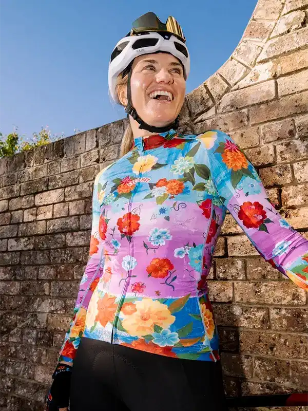 Woman in colorful floral cycling jersey and helmet, smiling outdoors near brick wall