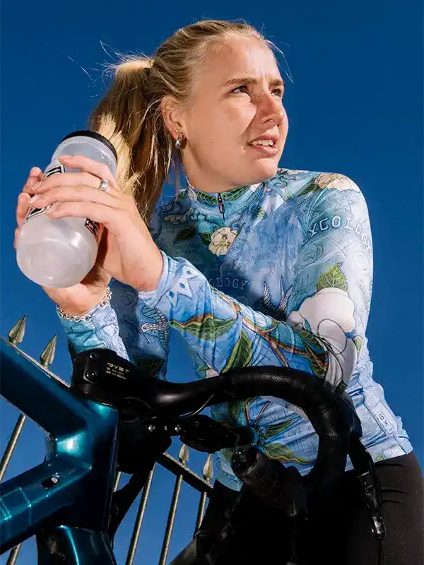 Woman in floral cycling jersey holding water bottle, riding bike under blue sky