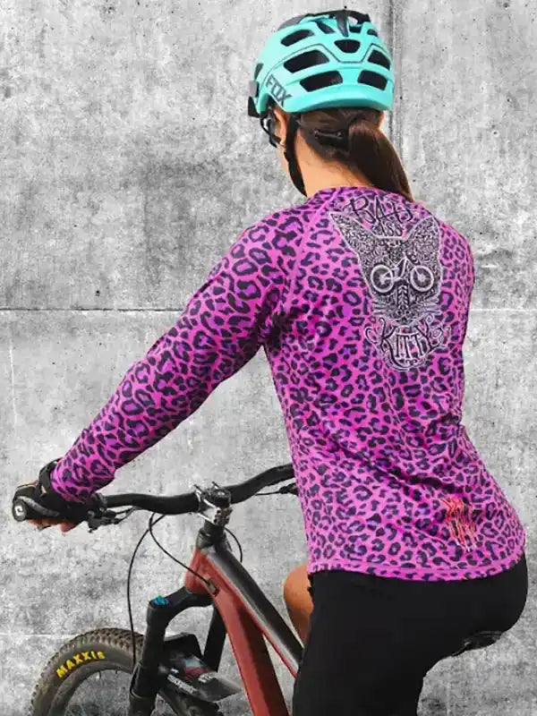 Woman cycling in vibrant pink leopard print long sleeve jersey with owl graphic, wearing teal helmet