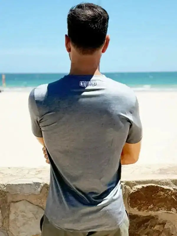 Man in gray cycling jersey with blue back panel, standing on beach, ocean view