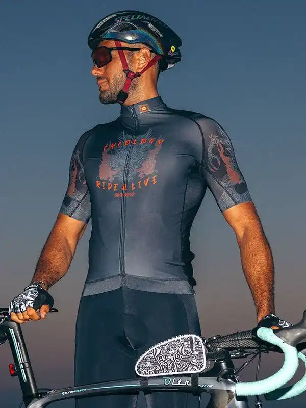 Man in black cycling jersey with 'Ride & Live' graphic, helmet, and gloves, standing with road bike at dusk