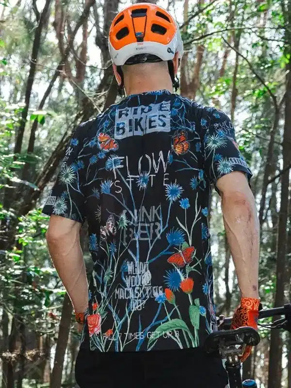 Man wearing floral cycling jersey with 'Ride Bikes' text, orange helmet, in forest trail