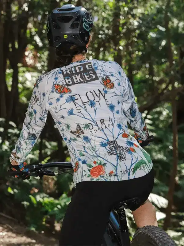 Woman cycling in floral long-sleeve jersey with 'Ride Bikes' print, forest trail, mountain bike gear
