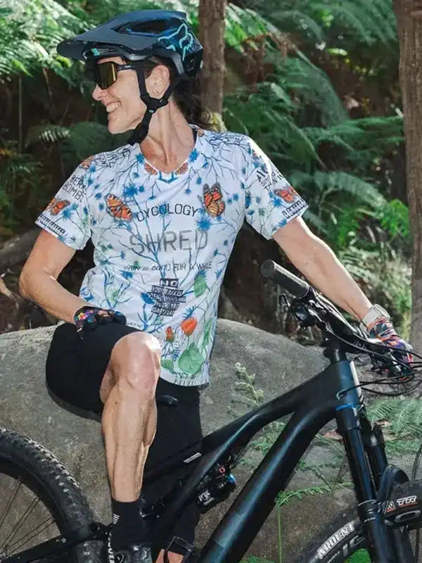 Woman in butterfly-print cycling jersey and helmet, smiling beside mountain bike in forest