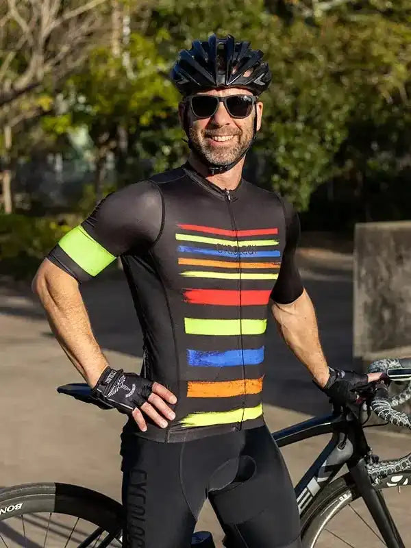 Man in colorful striped cycling jersey and black bib shorts, wearing helmet and sunglasses, standing with road bike outdoors