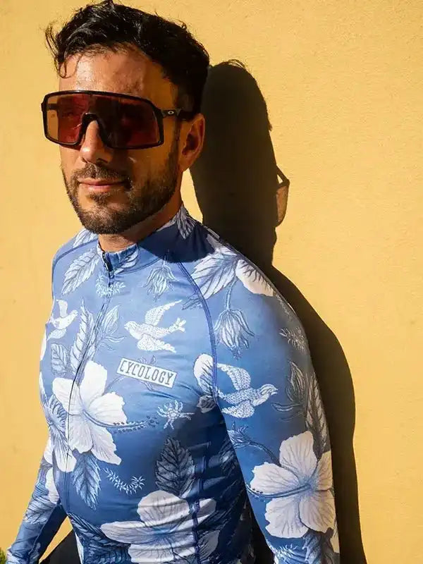 Man in blue floral cycling jersey with white hibiscus print, wearing sunglasses, against yellow wall
