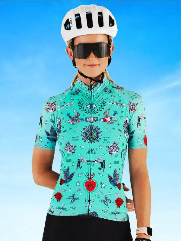 Woman in turquoise tattoo-style cycling jersey with birds, roses, and stars, wearing white helmet and sunglasses