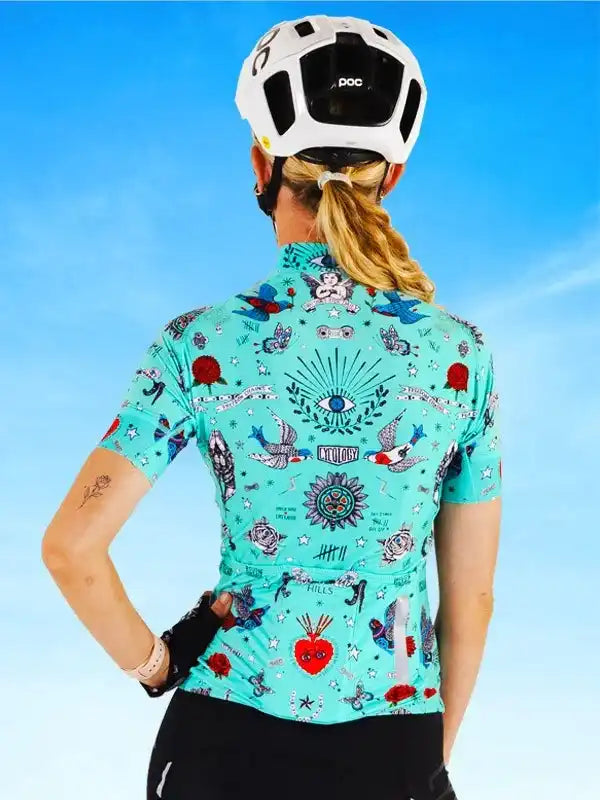Woman wearing turquoise tattoo-style cycling jersey with birds, hearts, and eye motifs, paired with POC helmet