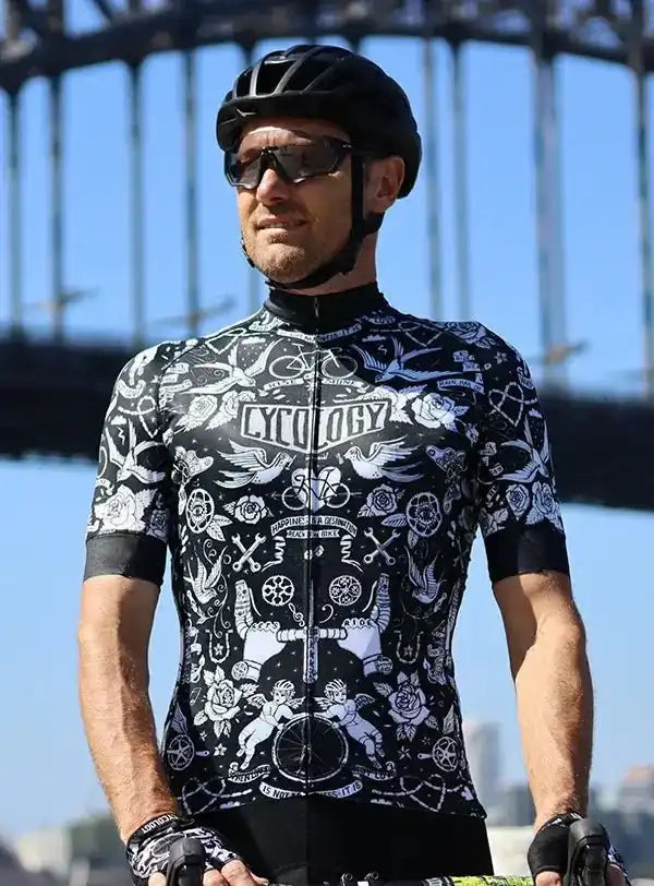 Man wearing black and white tattoo-style cycling jersey with 'Cycology' logo, helmet, and sunglasses outdoors
