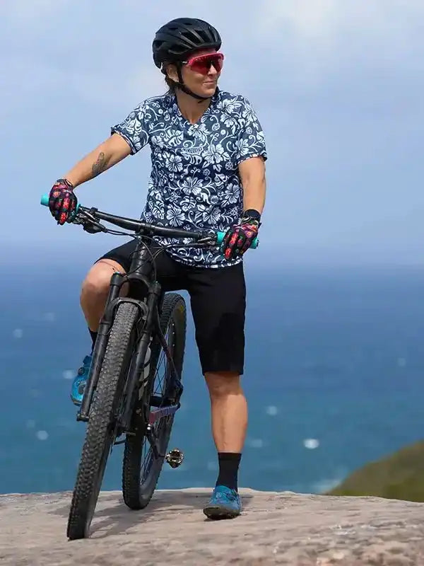 Cyclist in floral jersey and black shorts on mountain bike by ocean, wearing helmet and sunglasses