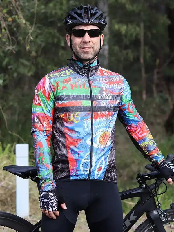 Man wearing colorful long-sleeve cycling jersey with graphic print, helmet, and gloves, standing beside road bike outdoors