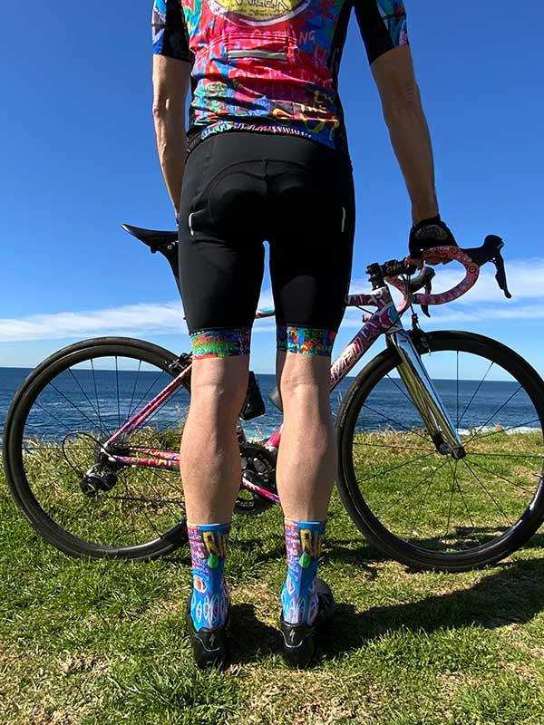 Cyclist in colorful cycling jersey and patterned socks standing with road bike by ocean, showcasing men's cycling apparel
