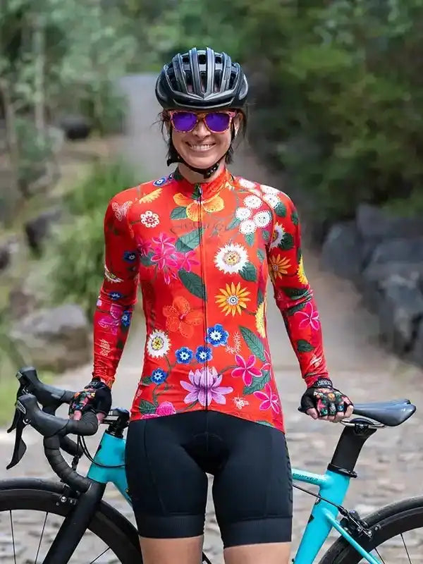 Woman in vibrant floral cycling jersey and black shorts, smiling with bike on forest trail