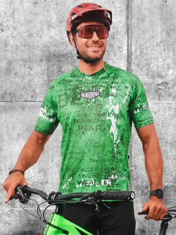 Man in green cycling jersey with graphic print, helmet, and sunglasses, holding bike against concrete wall