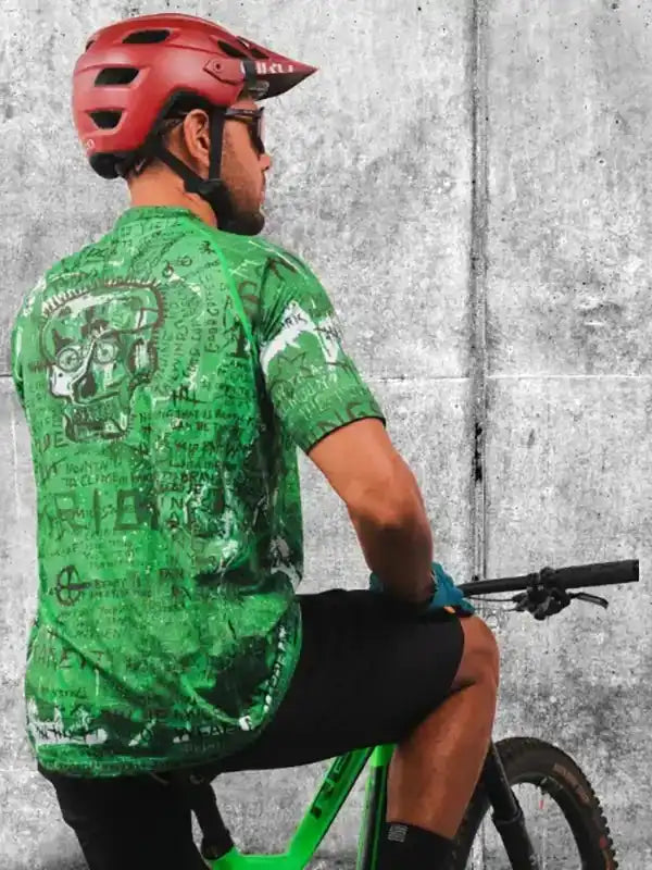 Man wearing green graphic cycling jersey and red helmet on mountain bike against concrete wall