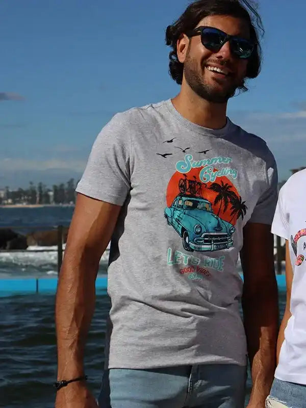 Man wearing gray Summer Cycling t-shirt with vintage car graphic, beach backdrop, perfect for casual rides.