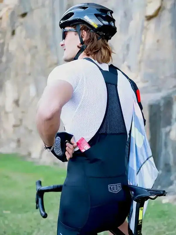 Man in black cycling bib shorts and white jersey, wearing helmet, holding bike outdoors
