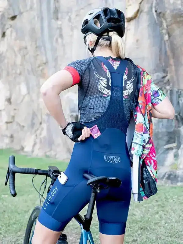 Woman in blue cycling bib shorts and jersey, standing with road bike near rocky cliff, wearing helmet and gloves