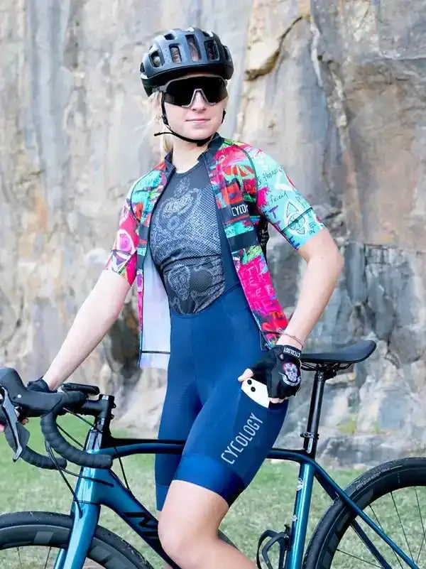 Woman in colorful cycling jersey and blue bib shorts with Cycology logo, standing with road bike outdoors